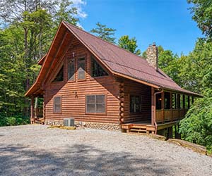 lodge on side of hill with wrap around deck. Tall wooded area around lodge.