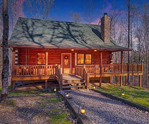 log cabin with green roof and front and side wrap deck and porch area on a wooded area